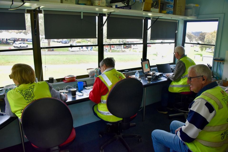 Volunteer officials in high-visibility MG Car Club Victoria vests working at desks in a race control room overlooking a motorsport venue