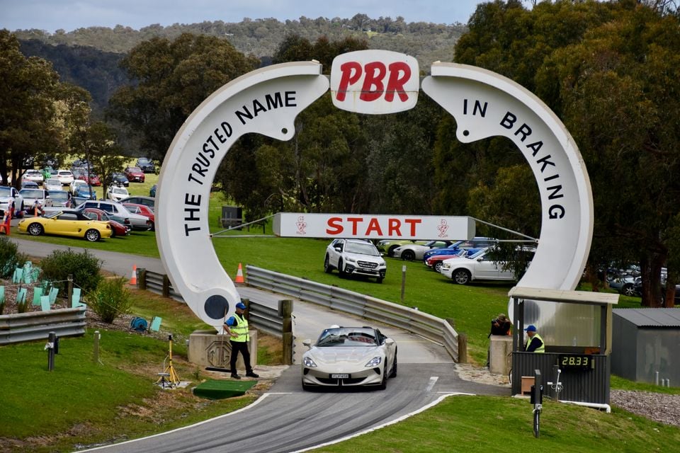 A white 2026 MG Cyberster convertible driving under the PBR start-line arch at a hillclimb event, with marshals and parked cars nearby