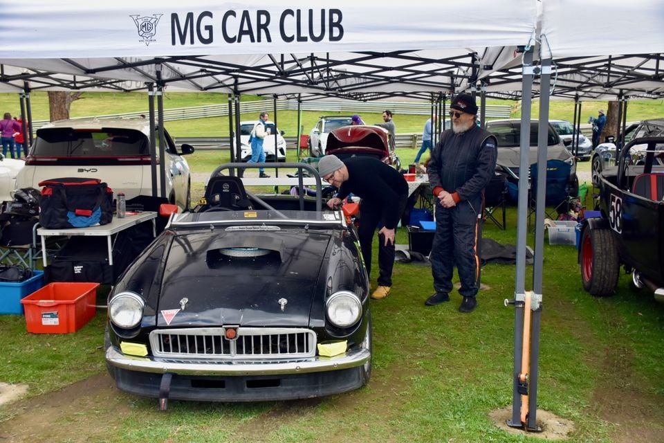 A black classic MG roadster race car parked under an MG Car Club marquee at a motorsport event, with two men looking at it