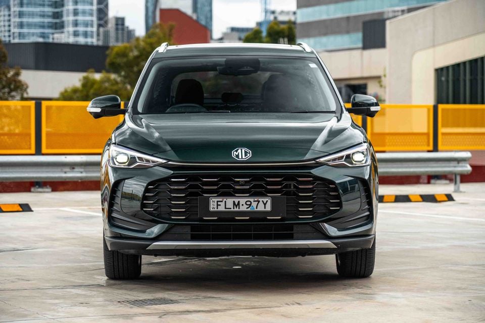 A dark green MG ZS SUV seen from the front, parked in an urban car park with city buildings in the background