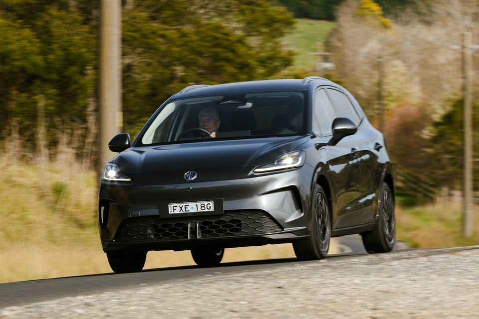 A dark grey 2026 MG4 Urban EV hatchback driving on a country road, seen from the front