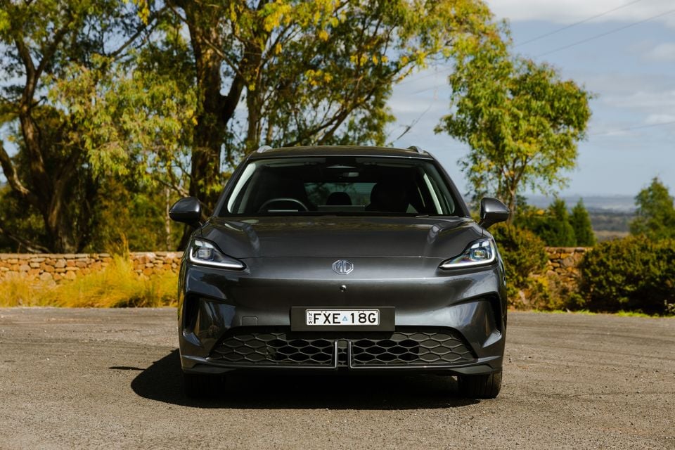 A grey MG4 electric hatchback seen from the front, parked on a gravel surface with trees and a stone wall in the background.