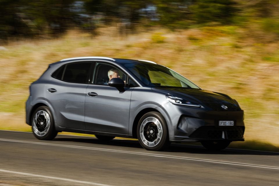 A grey 2026 MG4 Urban EV hatchback driving on a country road, seen from the front-side