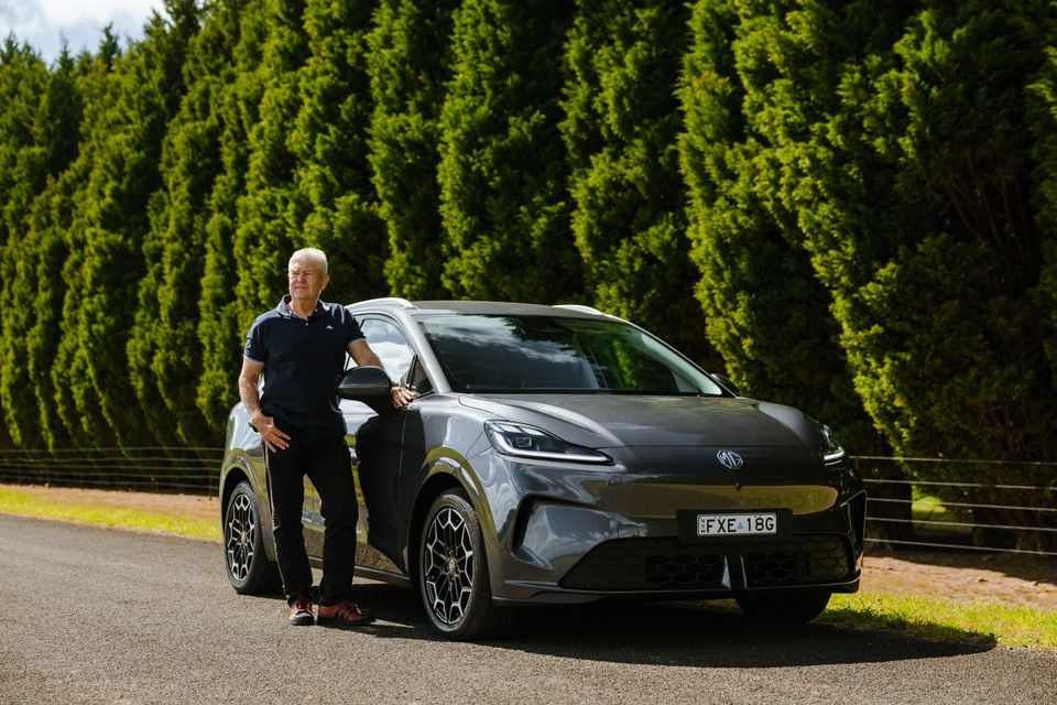 A man standing beside a grey MG4 Urban EV hatchback on a rural road, with tall hedges in the background
