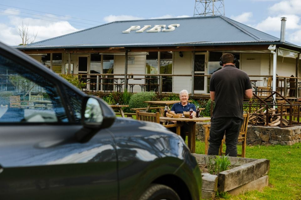 A person being filmed sitting at an outdoor table in front of a country pie shop, with a dark-coloured car in the foreground