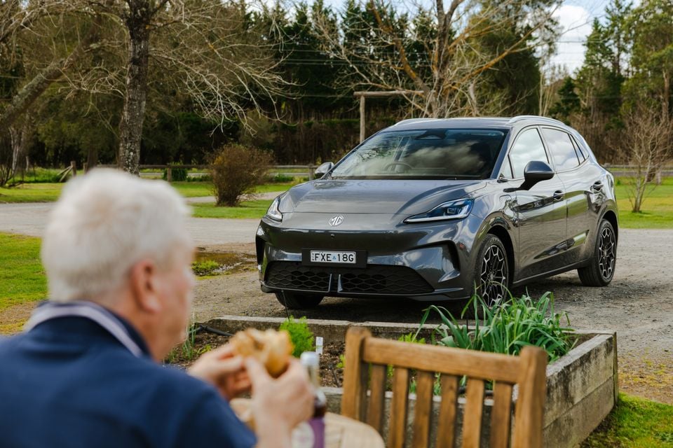 A grey MG4 electric SUV parked on gravel at a rural property, seen from the front, with a person eating in the foreground