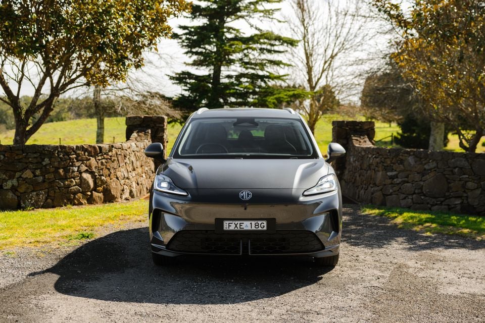 A grey MG4 SUV seen from the front, parked on a gravel driveway beside a stone wall in a rural setting.