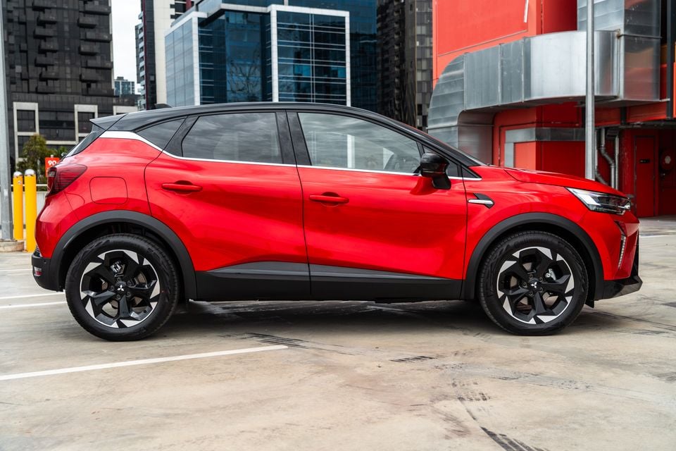 A red 2026 Mitsubishi ASX SUV seen from the side, parked in an urban car park with city buildings in the background
