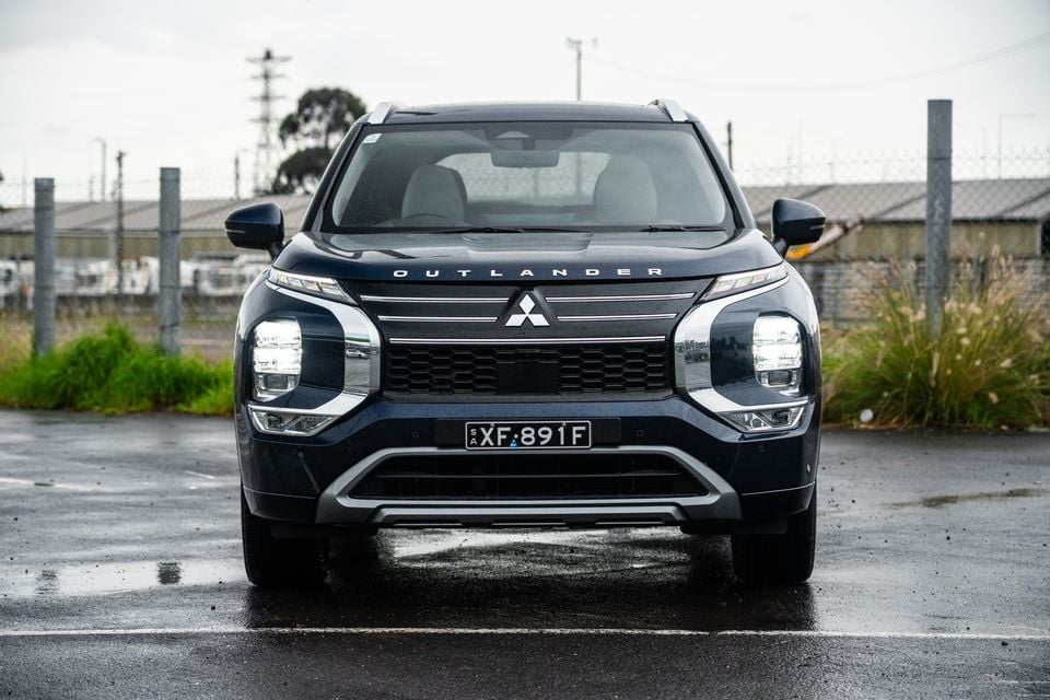 A dark blue Mitsubishi Outlander SUV seen from the front, parked on wet pavement on an overcast day.