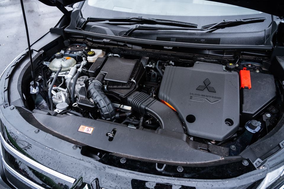 The open engine bay of a Mitsubishi Outlander Plug-In Hybrid EV, showing the PHEV motor cover with Mitsubishi logo and orange high-voltage wiring