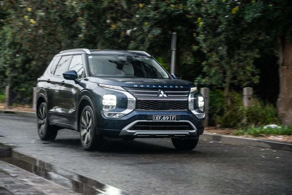 A dark blue Mitsubishi Outlander SUV driving on a wet road surrounded by trees
