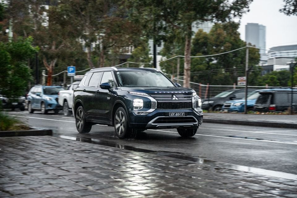 A dark blue Mitsubishi Outlander PHEV SUV driving on a wet city street in Australia, seen from the front