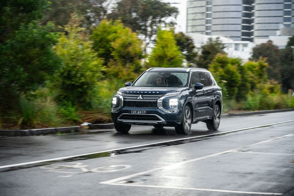 A dark blue Mitsubishi Outlander SUV driving on a wet road in rainy conditions, seen from the front
