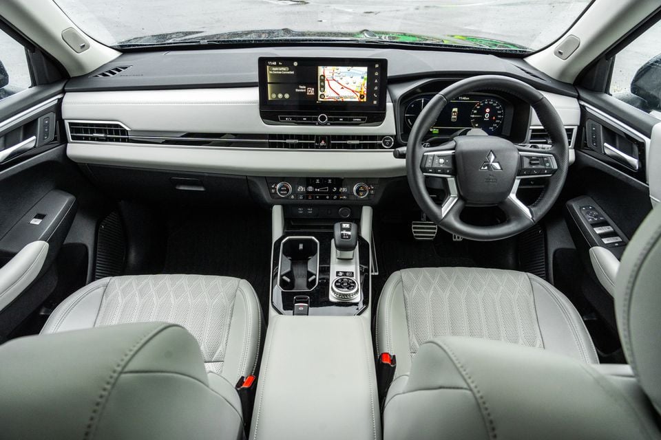 Interior of a 2026 Mitsubishi Outlander PHEV showing the dashboard, steering wheel, centre console, infotainment screen and light grey leather seats from the rear seats looking forward
