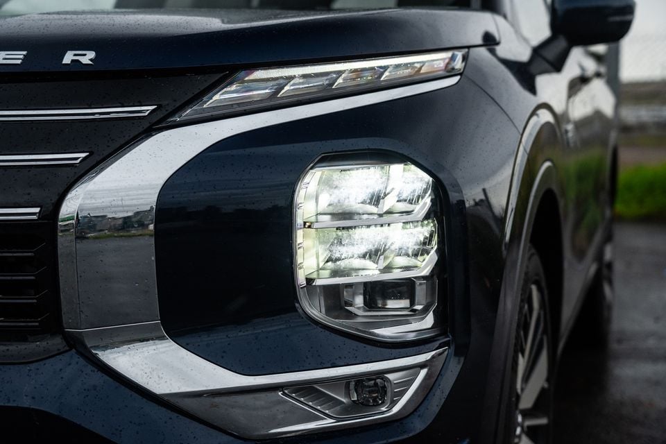 Close-up of the LED headlight on a dark blue 2026 Mitsubishi Outlander PHEV, showing the distinctive square headlight cluster and DRL strip