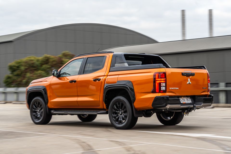 An orange 2026 Mitsubishi Triton GSR dual-cab ute driving across a rooftop car park, seen from the rear and driver's side