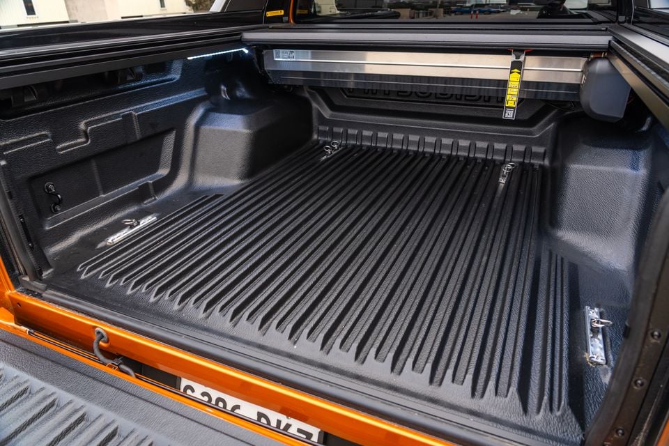 The tub of an orange 2026 Mitsubishi Triton GSR ute with a black bedliner, tie-down points and a roller tonneau cover, seen over the open tailgate