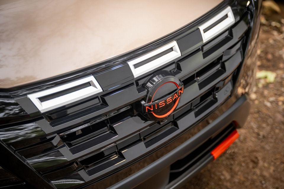 Close-up of the Nissan badge on the front grille of a 2026 Nissan Rogue Rock Creek Edition, featuring an orange-ringed emblem and silver accent trim.