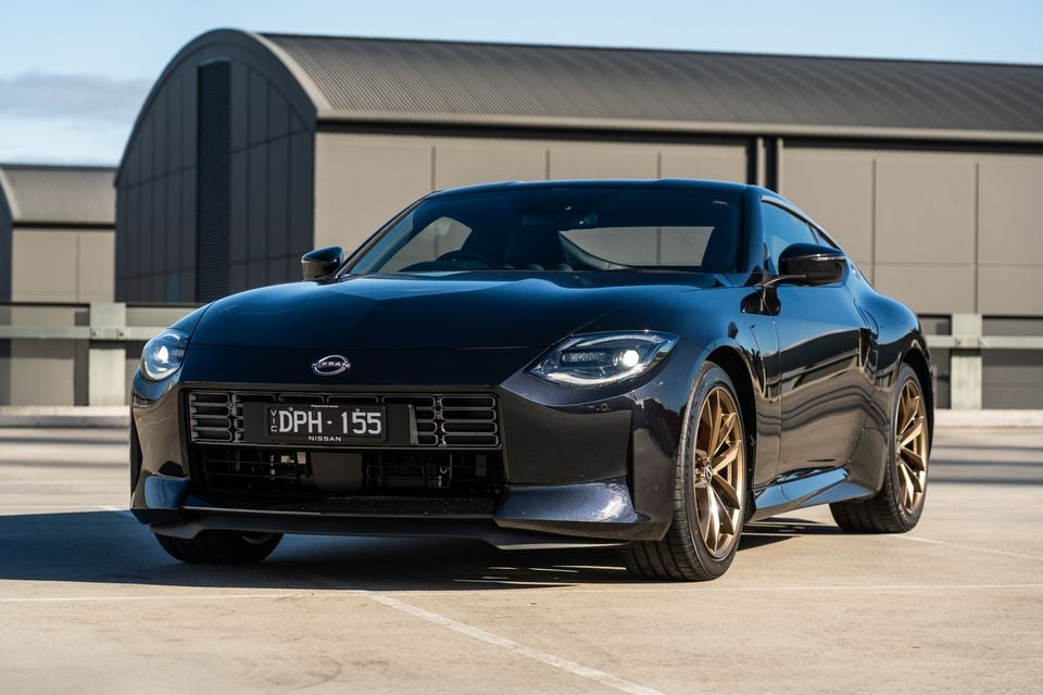 A dark blue 2026 Nissan Z sports coupe seen from the front, with gold alloy wheels, parked on a concrete surface near a large industrial shed.