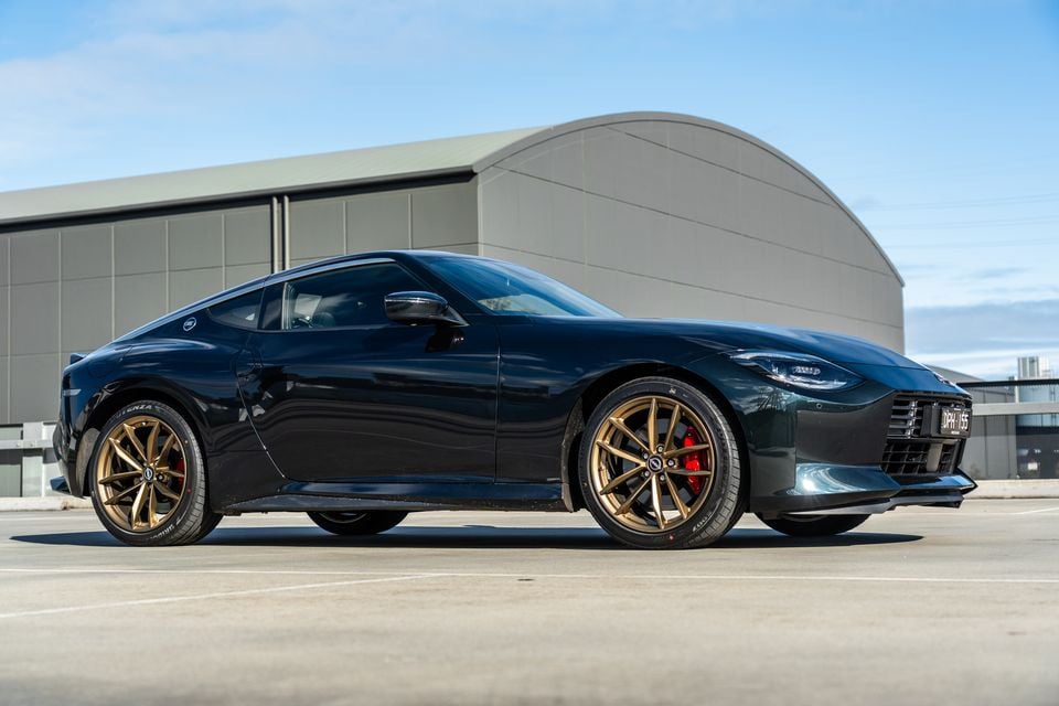 A dark green Nissan Z coupe with gold wheels and red brake calipers, seen from the front-side angle in a car park near a large industrial building