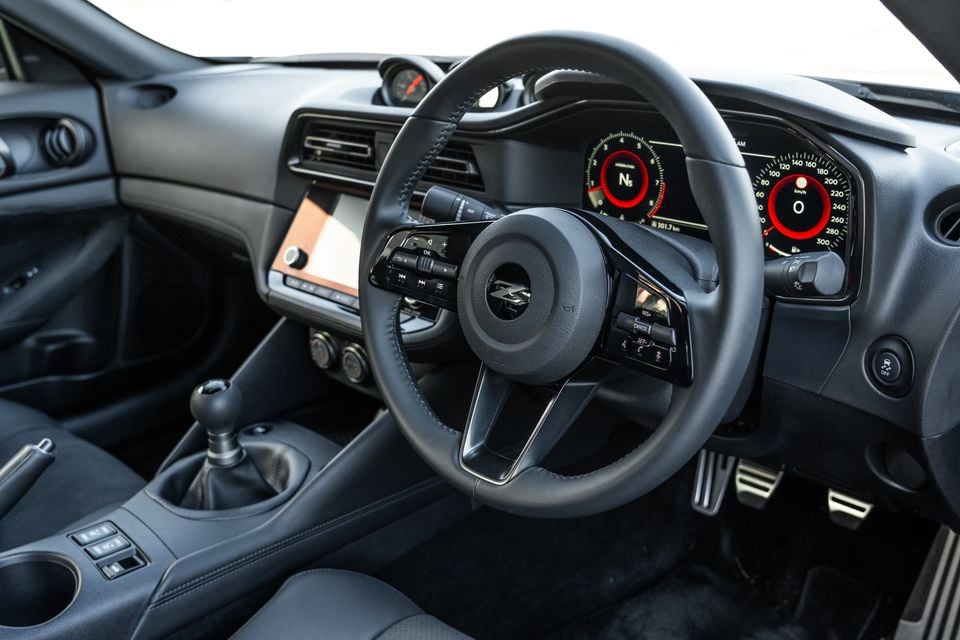 Interior of a Nissan Z showing the steering wheel with Z badge, manual gear shifter, digital instrument cluster with red-accented gauges, and infotainment touchscreen