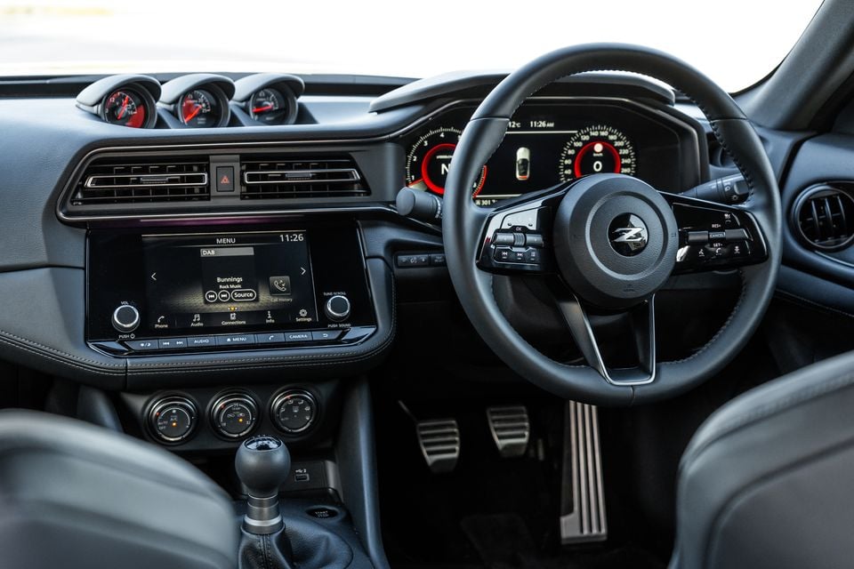 Interior of a Nissan Z showing the dashboard, steering wheel with Z badge, infotainment screen, manual gear shifter, triple auxiliary gauges on the dash top, and digital instrument cluster