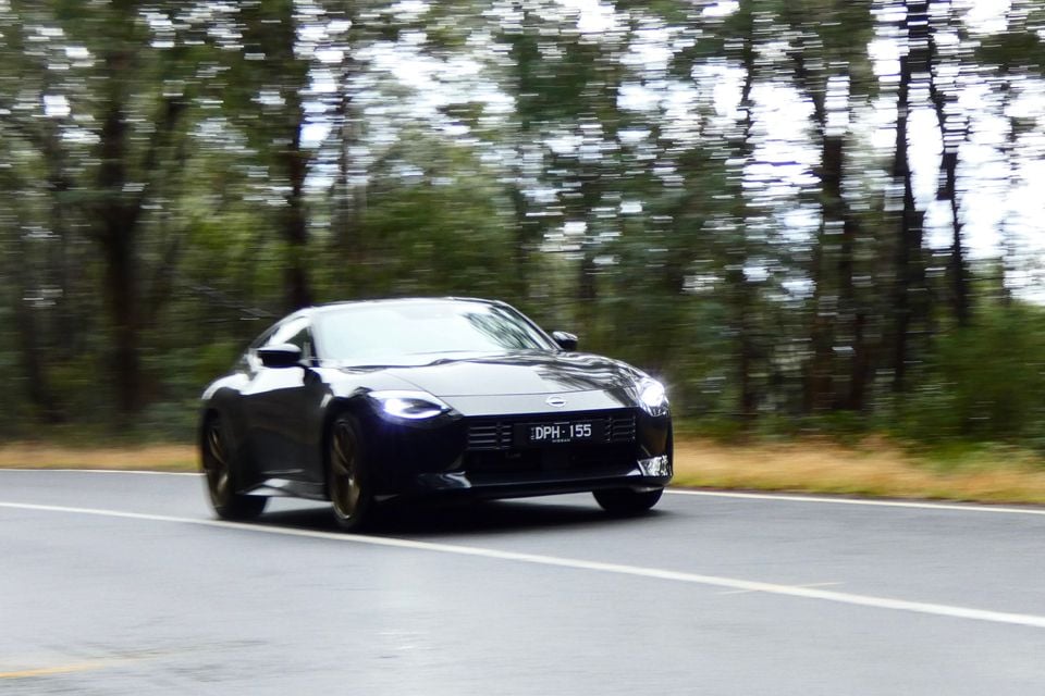 A dark blue Nissan Z coupe driving along a country road, seen from the front, with trees blurred in the background