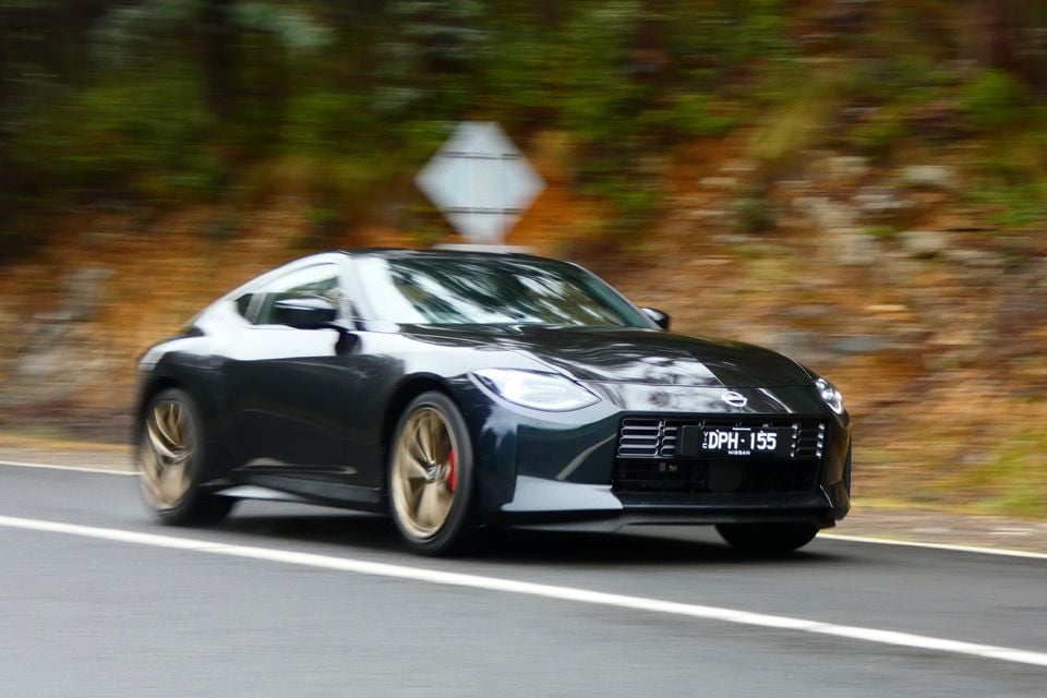 A dark blue 2026 Nissan Z coupe seen from the front-side angle driving on a road, with gold wheels and red brake calipers visible