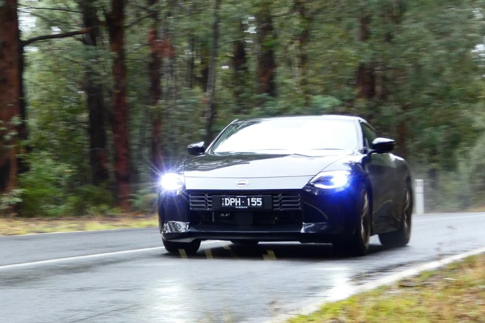 A dark blue Nissan Z coupe driving towards the camera on a wet forest road with headlights on