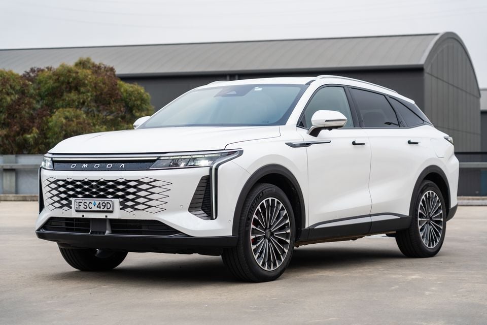A white Omoda 9 SUV seen from the front-side angle, parked on a concrete surface in front of an industrial building.