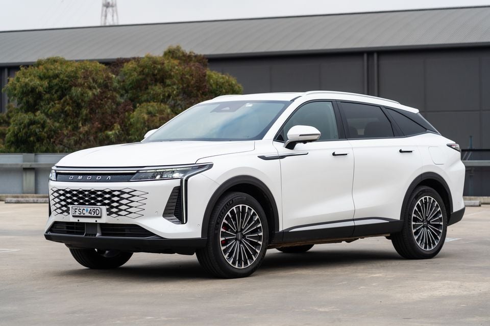 Um SUV Omoda 9 branco visto pela frente esquerda, estacionado em uma área aberta com um prédio e árvores ao fundo.