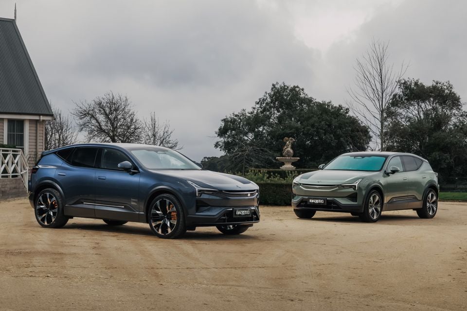 Two Polestar electric SUVs parked side by side on a gravel driveway — a dark blue Polestar 3 on the left and a green Polestar 3 on the right — with a heritage building and garden statue in the background