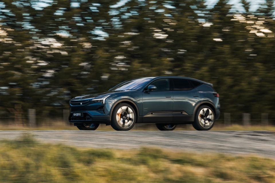 A teal 2026 Polestar 3 SUV seen from the side while driving on a rural road, with motion-blurred trees in the background
