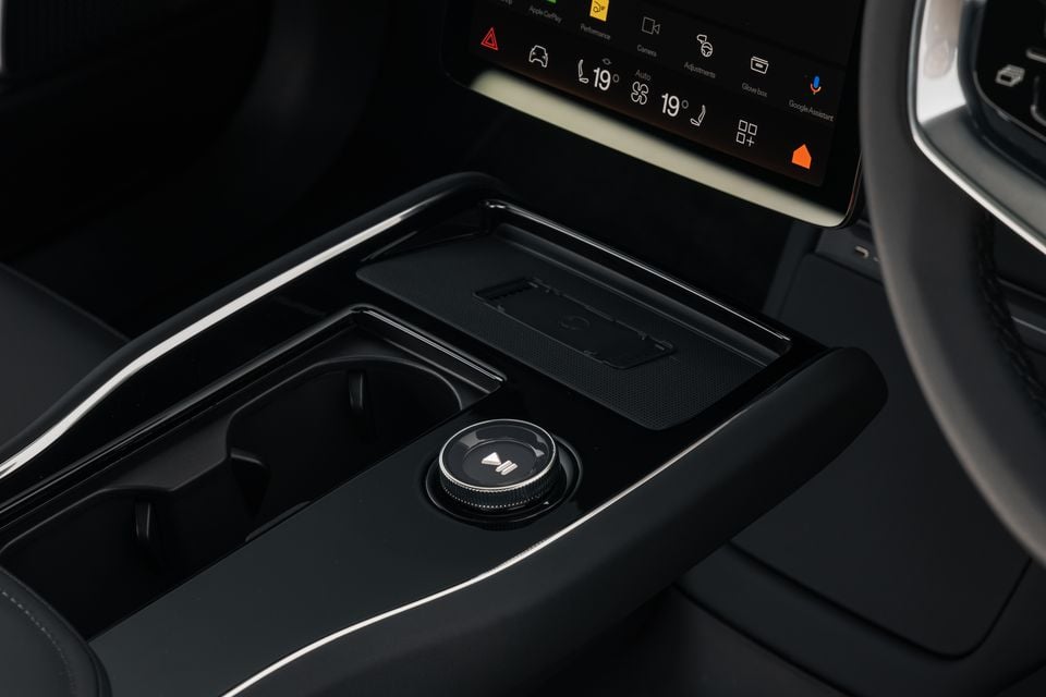Interior centre console of the 2026 Polestar 3 showing a rotary media control knob, wireless charging pad, cup holder, and touchscreen infotainment display with Apple CarPlay and Google Assistant icons visible.