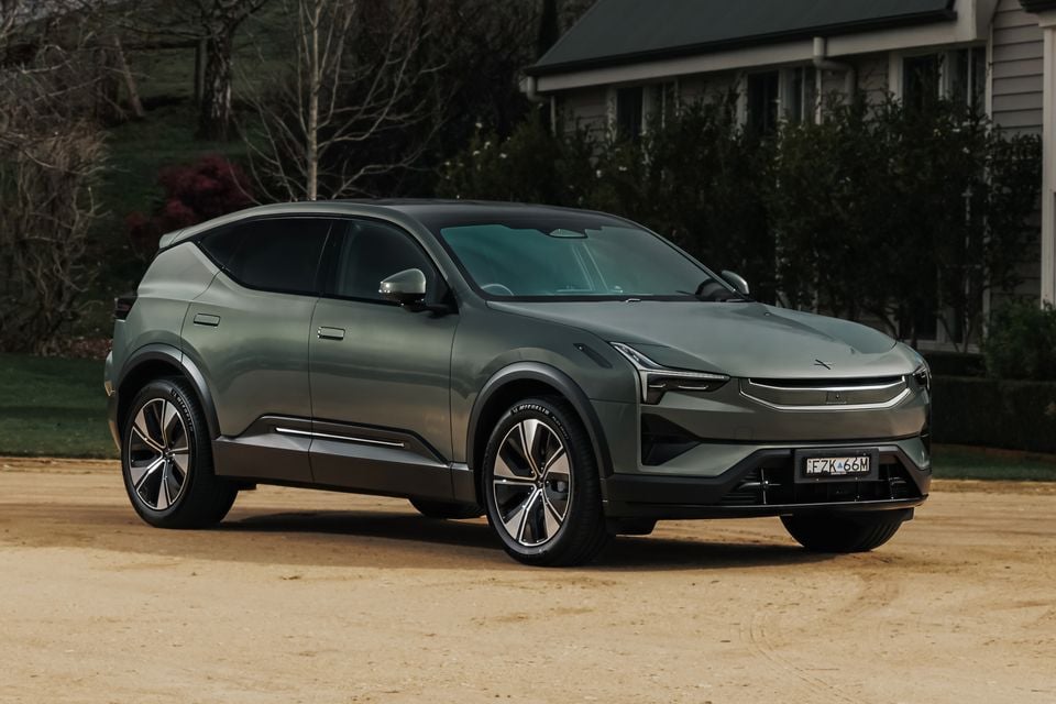 A grey 2026 Polestar 3 electric SUV seen from the front-left side, parked on a gravel driveway with a house and trees in the background