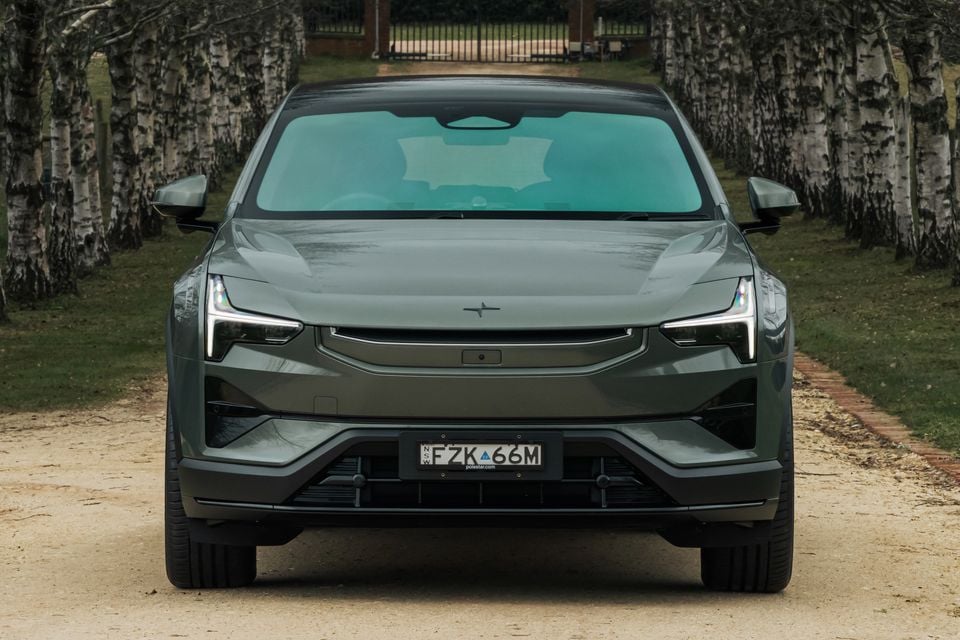 A dark green Polestar 3 SUV seen from the front, parked on a gravel driveway lined with birch trees.