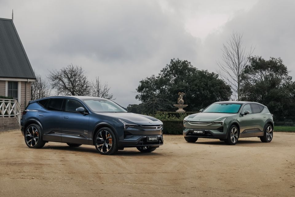 Two Polestar 3 electric SUVs parked on a gravel driveway near a country estate — one in dark blue and one in green — seen from the front and side