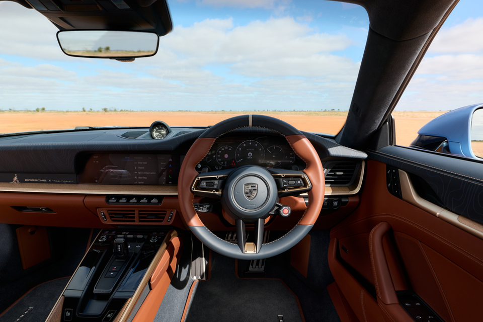 Interior of a Porsche 911 Turbo S showing the tan leather steering wheel, digital instrument cluster, touchscreen infotainment display, and centre console, shot from the driver's perspective with an arid outback landscape visible through the windscreen.