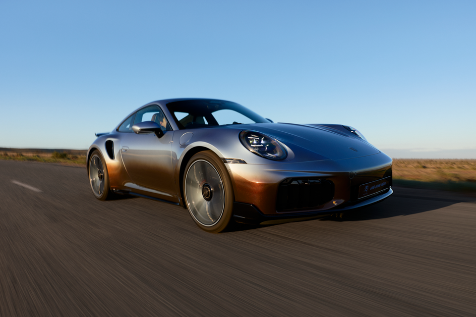 A Porsche 911 Turbo S coupe with a grey-to-bronze gradient finish driving on an open road at golden hour, seen from the front-side