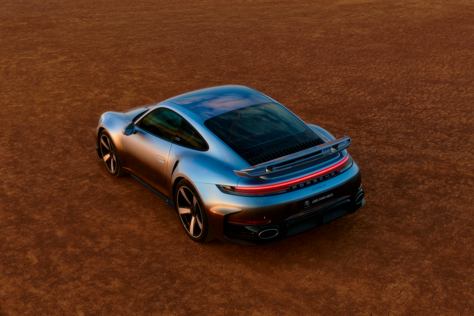 A silver Porsche 911 Turbo S coupe seen from a high rear three-quarter angle, parked on dry red earth terrain