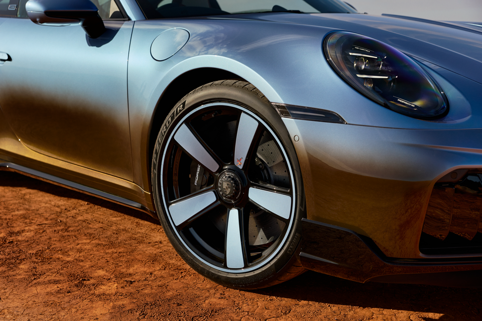 Close-up of the front wheel, alloy rim, and headlight of a Porsche 911 Turbo S in a metallic grey-gold finish, parked on red dirt ground