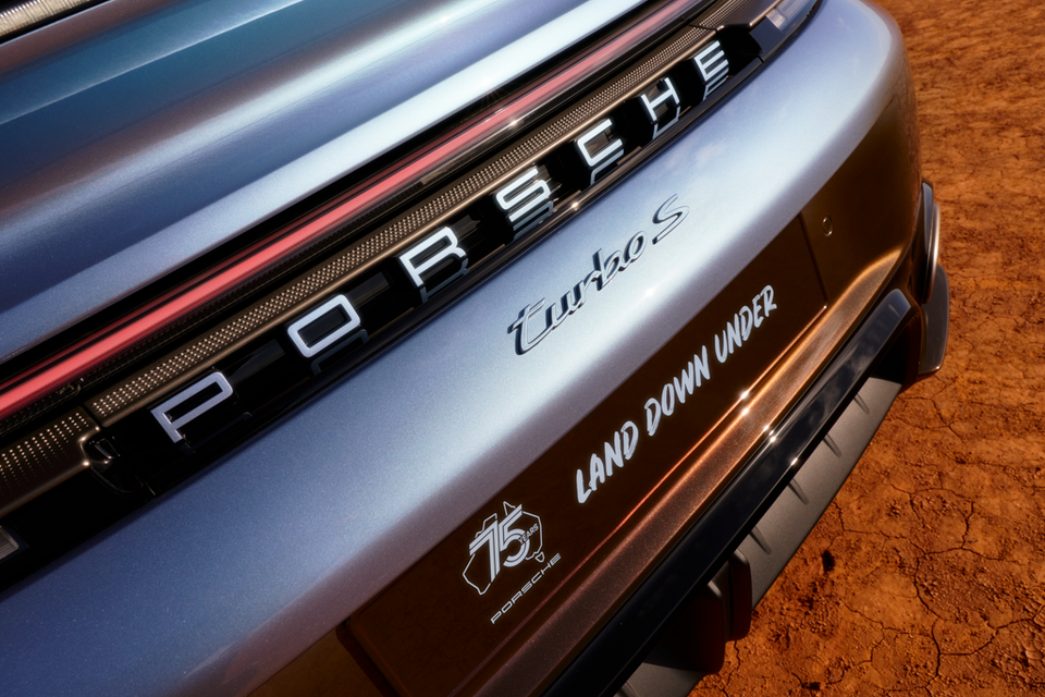 Close-up of the rear of a Porsche 911 Turbo S 'Land Down Under' special edition, showing the Porsche lettering, Turbo S badge, and 75 Years Porsche anniversary logo on the bumper, set against red Australian outback dirt.