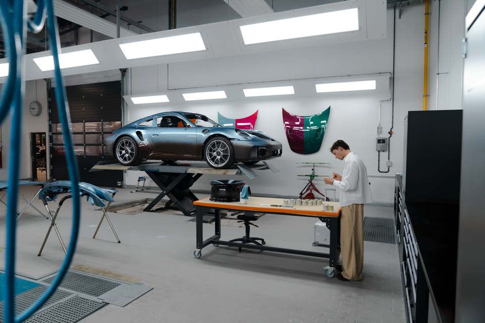 A grey Porsche 911 raised on a scissor lift inside a custom paint shop, with colourful painted body panel samples mounted on the wall and a technician working at a nearby workbench