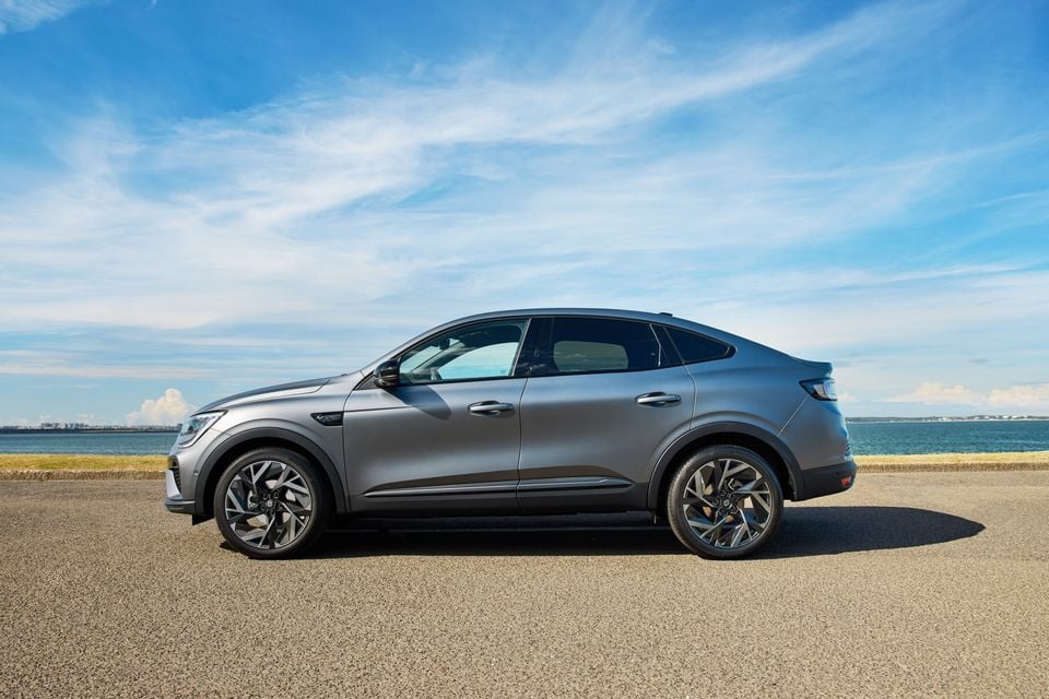 A grey 2026 Renault Arkana coupe-SUV seen from the side, parked near a waterfront under a blue sky.
