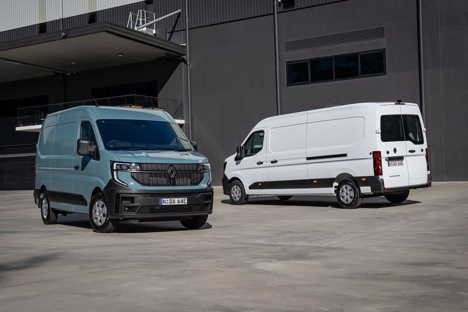 Two Renault Master vans parked outside a warehouse — a light blue example seen from the front and a white example seen from the rear