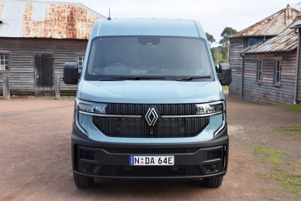 A light blue 2026 Renault Master van seen from the front, parked on a gravel surface beside rustic wooden buildings