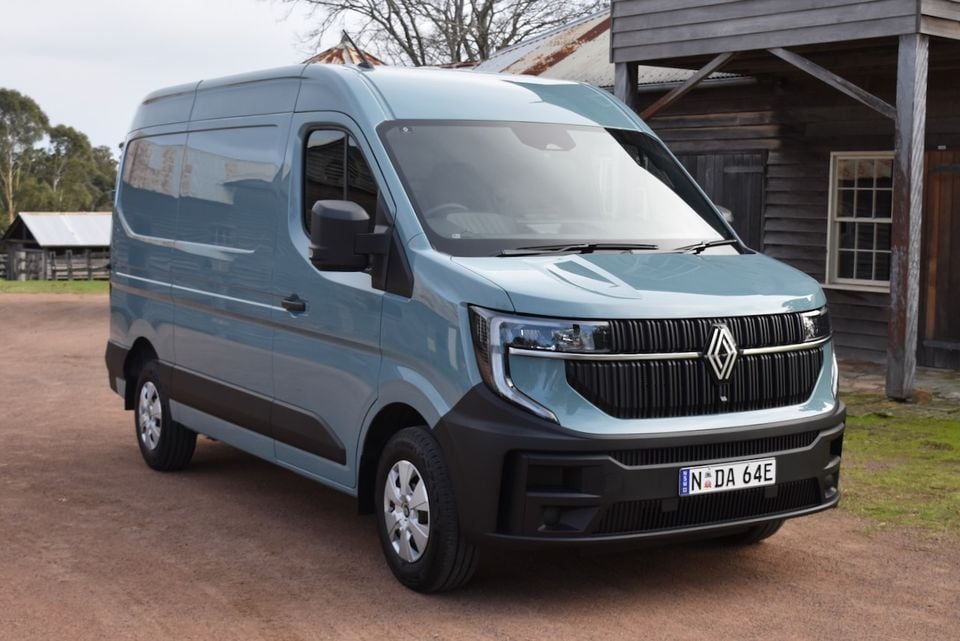 A blue 2026 Renault Master van seen from the front-side angle, parked on a gravel surface beside a rustic wooden building.