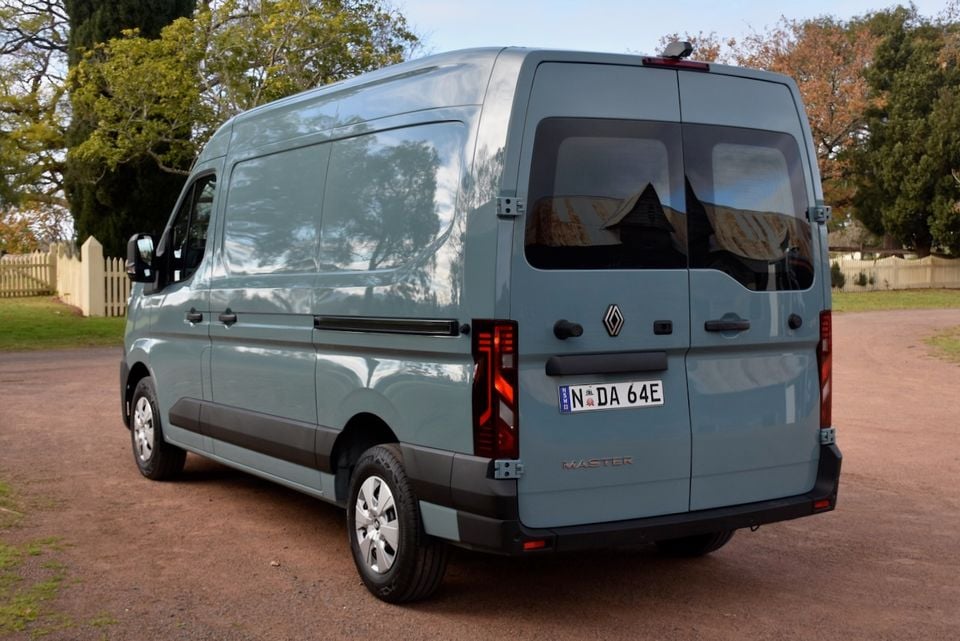 A blue-grey 2026 Renault Master van seen from the rear three-quarter angle, parked on a gravel driveway surrounded by trees