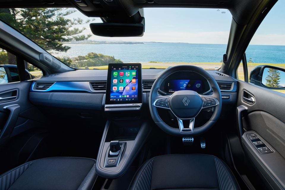 Interior of a Renault Symbioz Esprit Alpine showing the dashboard, large portrait touchscreen infotainment display running Apple CarPlay, digital instrument cluster, and Renault-badged steering wheel, with a coastal view through the windscreen