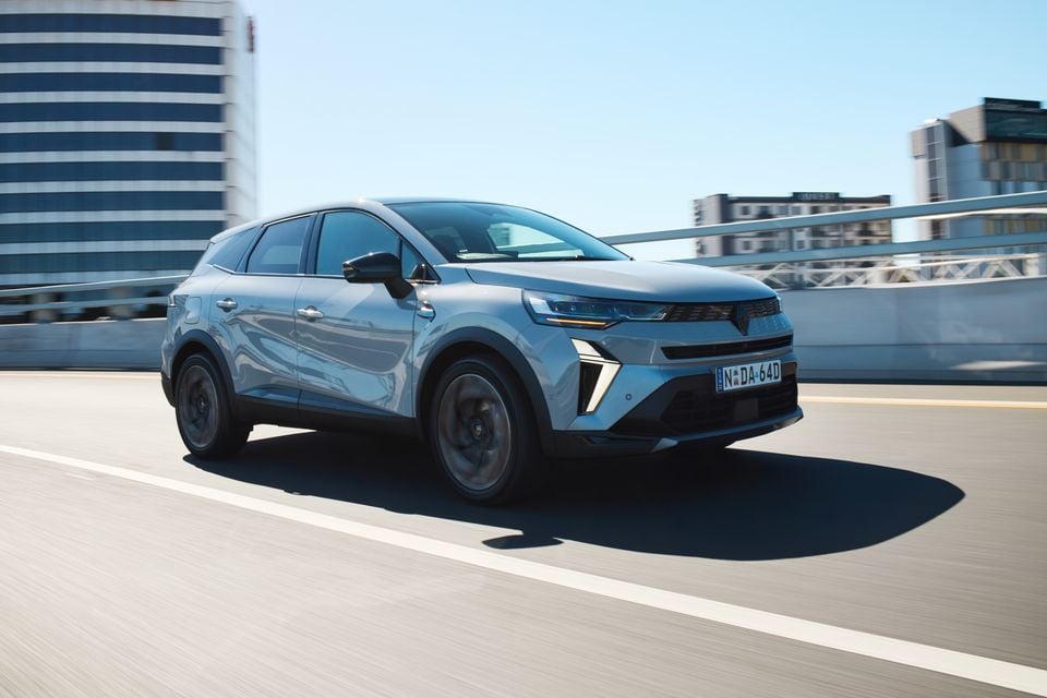 A grey 2026 Renault Symbioz Esprit Alpine SUV driving on a city road, seen from the front-side
